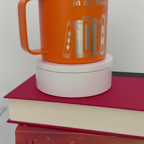 Orange stainless steel mug with 'in my bookish era' and books engraved around it, set on top of a stack of orange and red books.