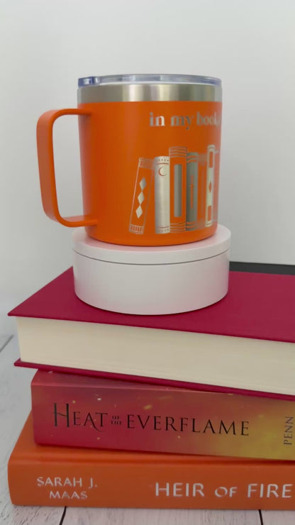 Orange stainless steel mug with 'in my bookish era' and books engraved around it, set on top of a stack of orange and red books.