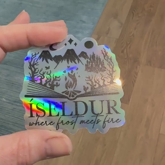 holographic sticker with an open book design and fantasy elements including mountains, fire, trees and castle and the words 'Iseldur: where frost meets fire' inspired by The Road of Bones by Demi Winters