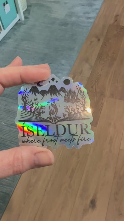 holographic sticker with an open book design and fantasy elements including mountains, fire, trees and castle and the words 'Iseldur: where frost meets fire' inspired by The Road of Bones by Demi Winters