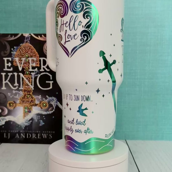 An Ever King themed white handled travel tumbler with a clear lid sits on a white pedestal in front of a teal wall and light wood floor. The tumbler is printed with the title 'The Ever: Beautiful Monster' in script and serif fonts, decorated with a serpent, ship, songbird and quotes in rainbow engraved details.