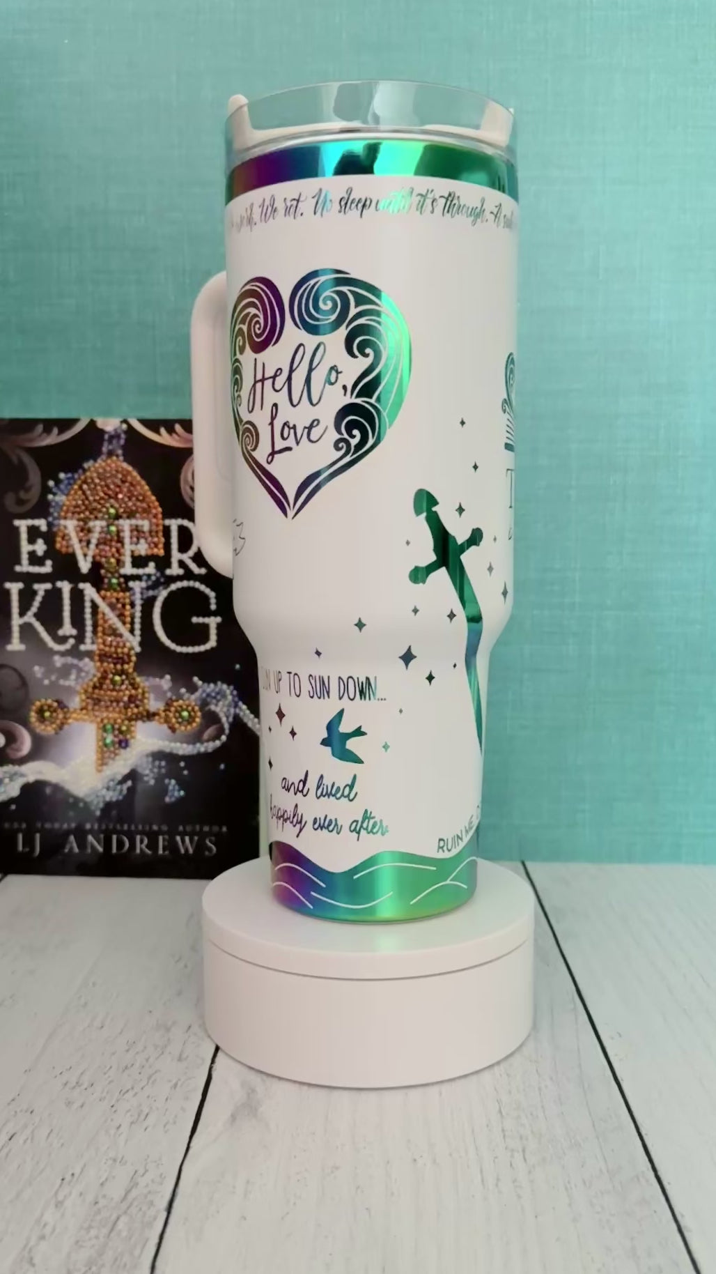 An Ever King themed white handled travel tumbler with a clear lid sits on a white pedestal in front of a teal wall and light wood floor. The tumbler is printed with the title 'The Ever: Beautiful Monster' in script and serif fonts, decorated with a serpent, ship, songbird and quotes in rainbow engraved details.