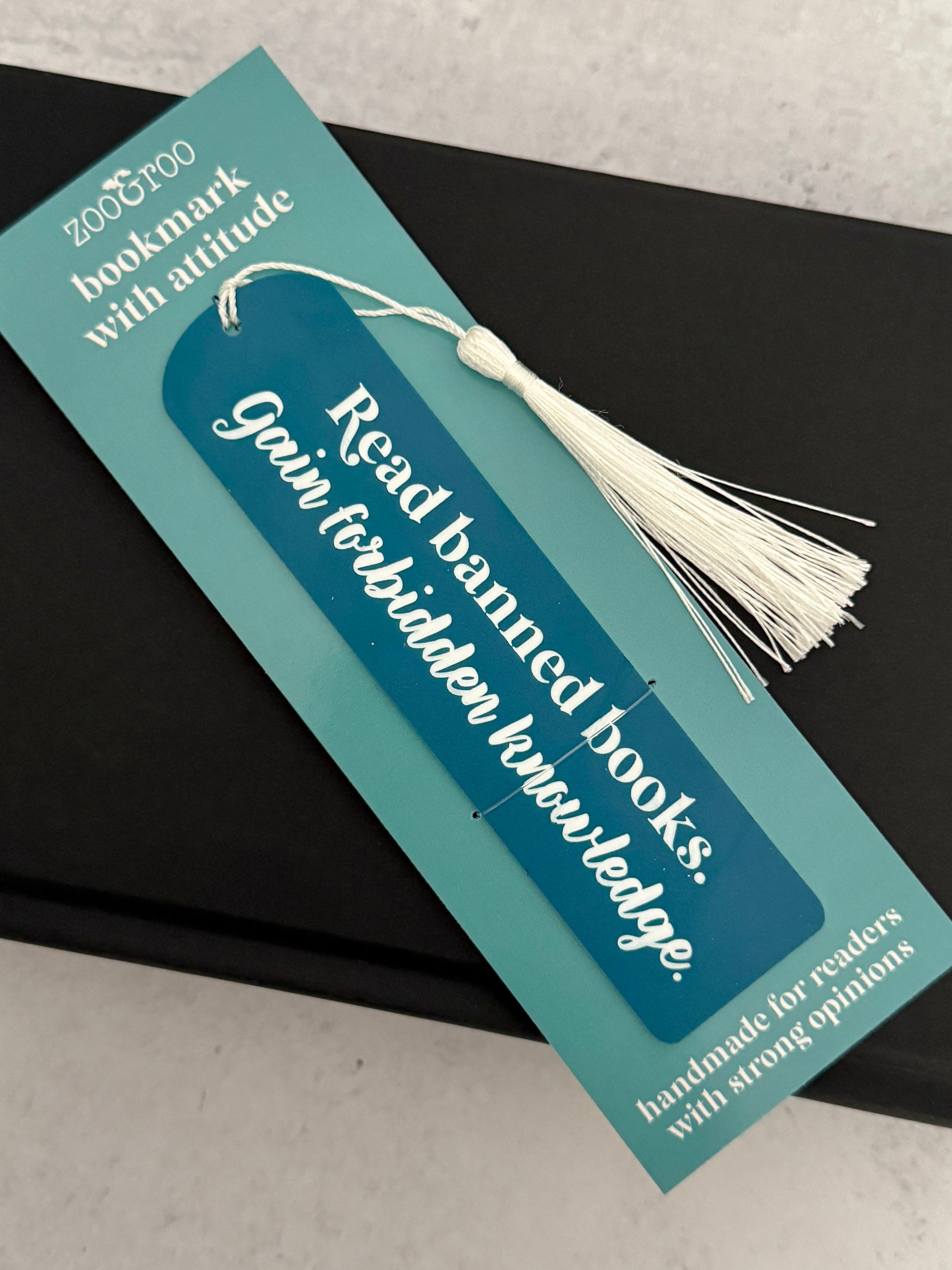 Bookish flatlay featuring slate blue acrylic banned books bookmark engraved with "read banned books, gain forbidden knowledge" in packaging that reads "bookmark with attitude" and "handmade for readers with strong opinions," resting on a black novel