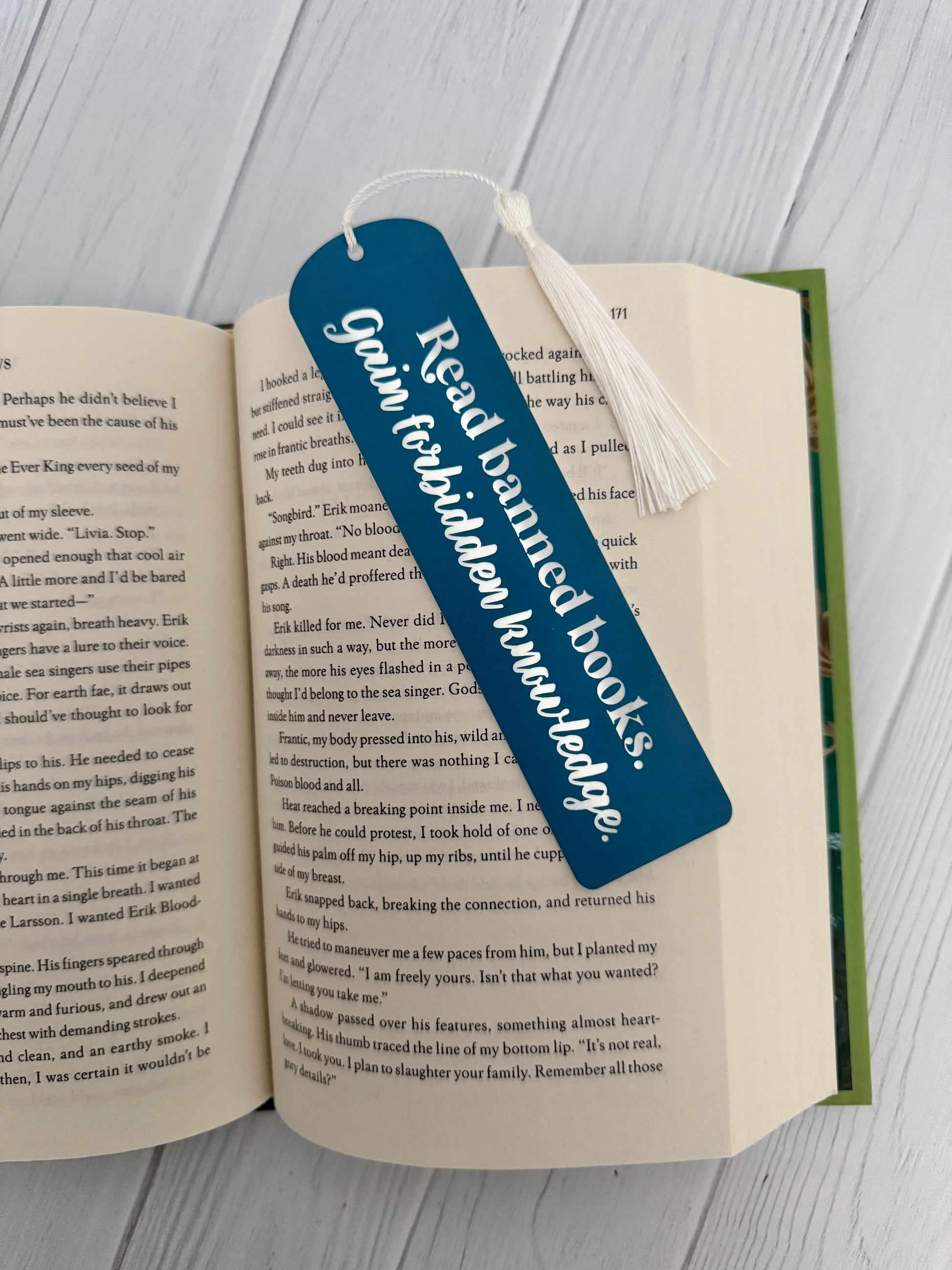 Slate blue acrylic bookmark engraved with “Read banned books, gain forbidden knowledge,” with white tassel, displayed on open book