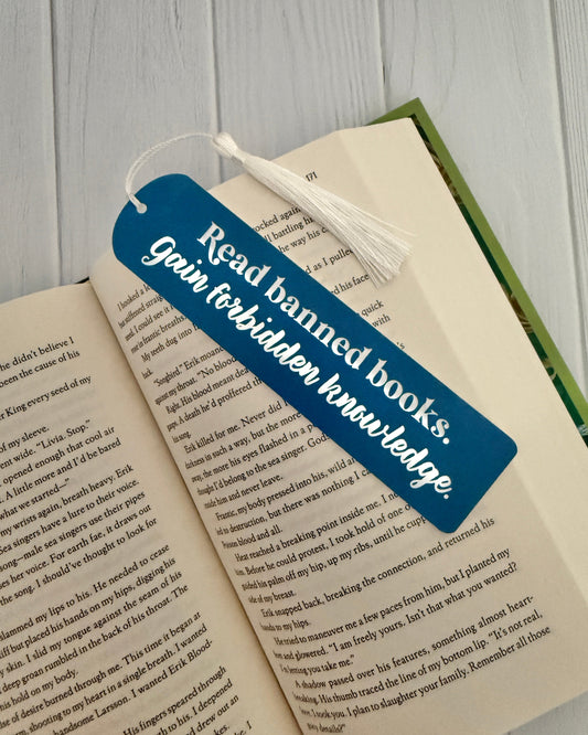 Slate blue acrylic bookmark engraved with “Read banned books, gain forbidden knowledge,” with white tassel, displayed on open book