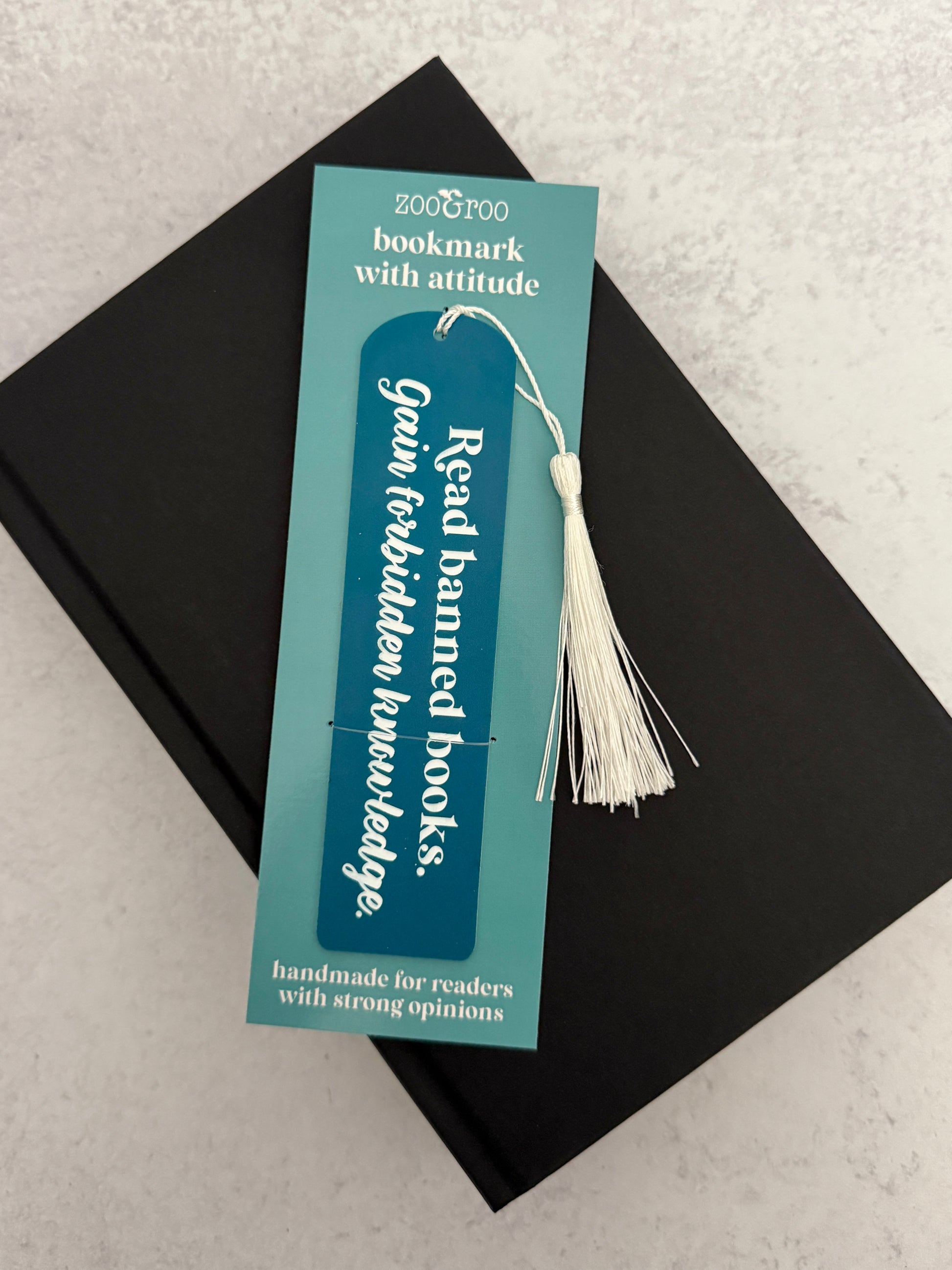 Bookish flatlay featuring slate blue acrylic banned books bookmark engraved with "read banned books, gain forbidden knowledge" resting on a black novel