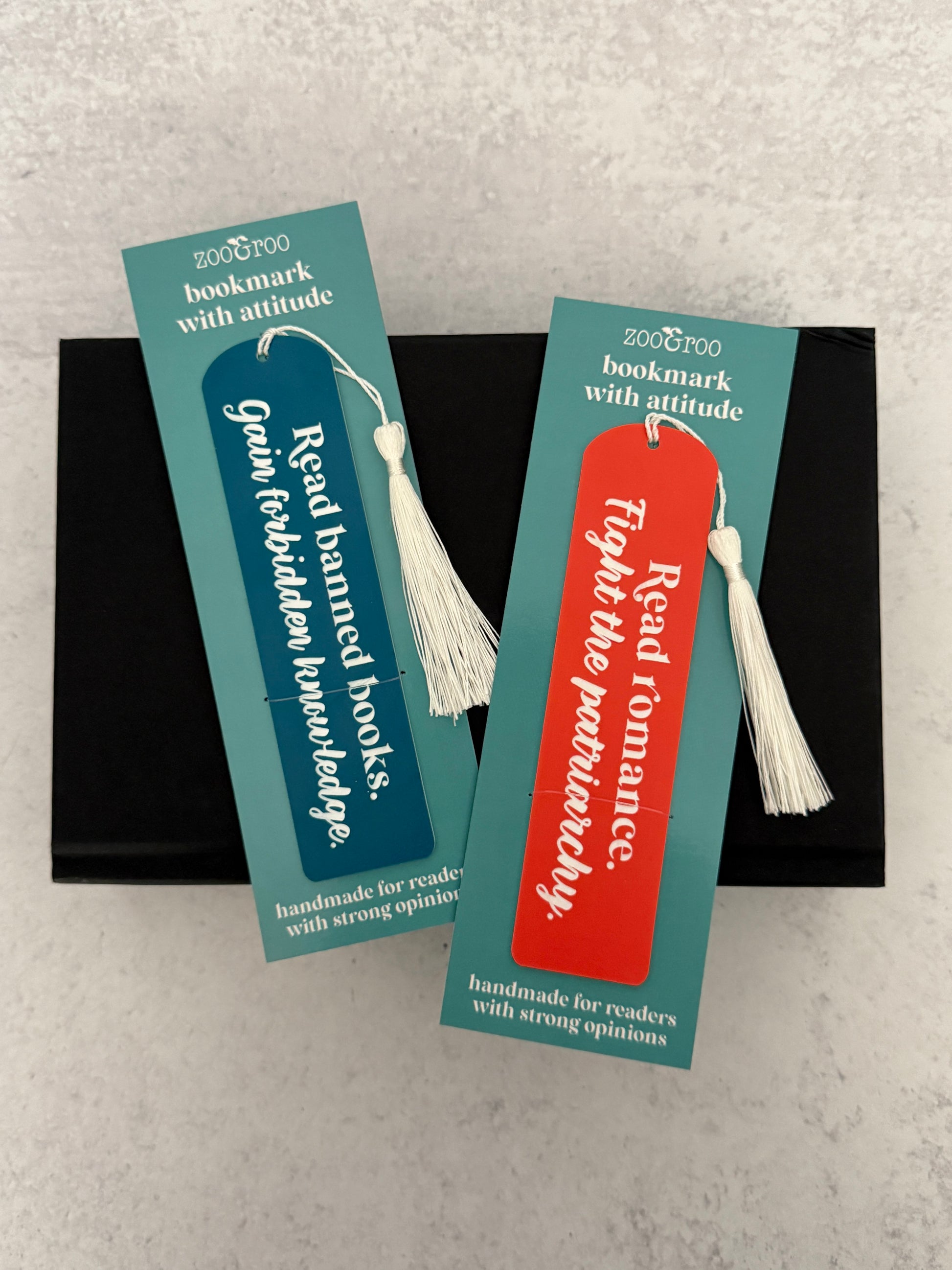 Two bookmarks engraved with "read banned books, gain forbidden knowledge"  and "read romance, fight the patriarchy" in packaging that reads "bookmark with attitude" and "handmade for readers with strong opinions," resting on a black novel