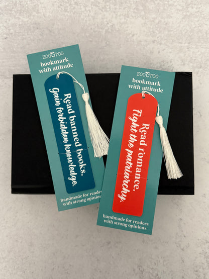 Two bookmarks engraved with "read banned books, gain forbidden knowledge"  and "read romance, fight the patriarchy" in packaging that reads "bookmark with attitude" and "handmade for readers with strong opinions," resting on a black novel