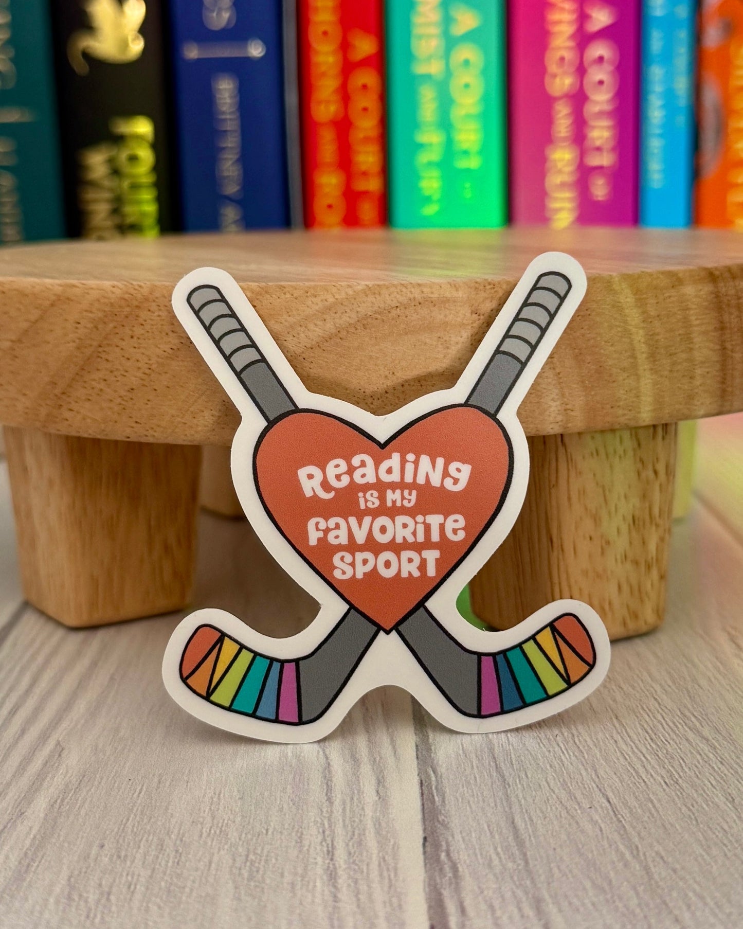 Sticker with 'Reading is my favorite sport' text in a heart with hockey sticks on a wooden block with books in the background