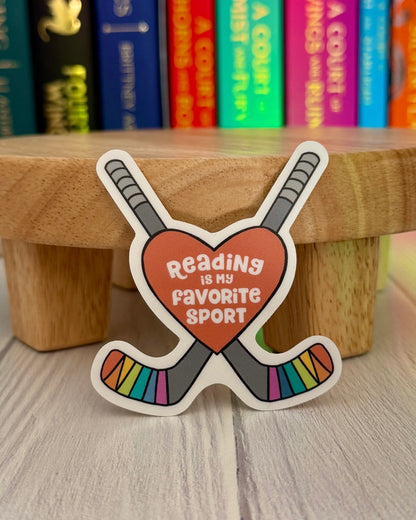 Sticker with 'Reading is my favorite sport' text in a heart with hockey sticks on a wooden block with books in the background