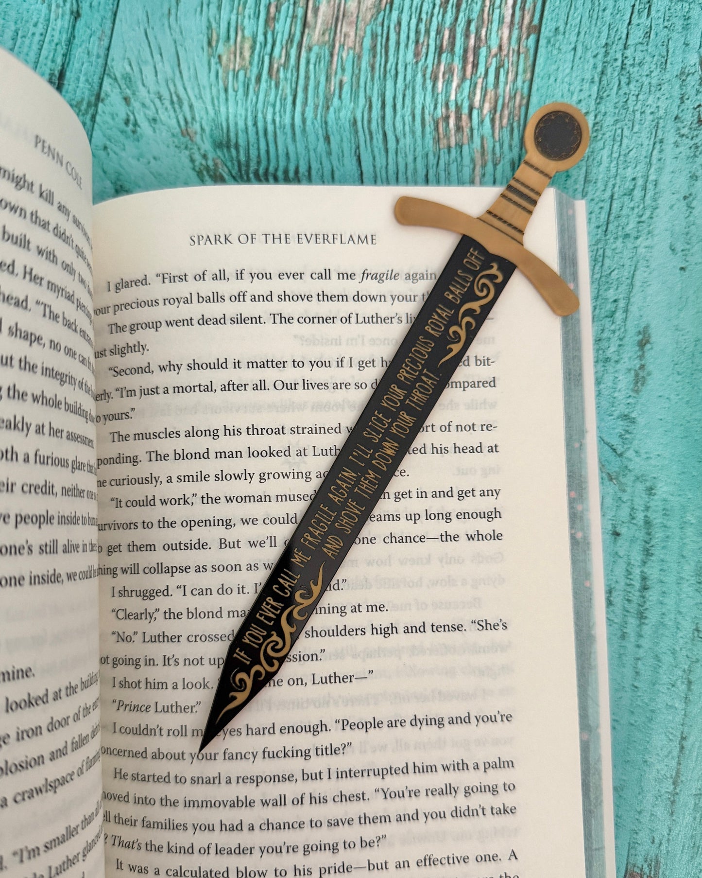 Decorative bookmark with sword design on an open book against a teal wooden background "If you ever call me fragile again, I'll slice your precious royal balls off and shove them down your throat." Officially licensed from The Kindred's Curse Saga by Penn Cole.