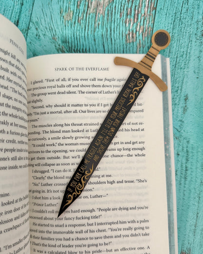 Decorative bookmark with sword design on an open book against a teal wooden background "If you ever call me fragile again, I'll slice your precious royal balls off and shove them down your throat." Officially licensed from The Kindred's Curse Saga by Penn Cole.
