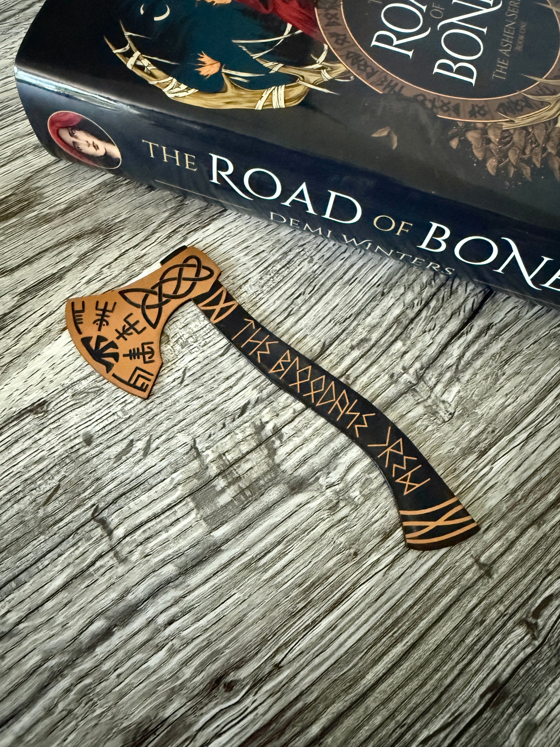 Decorative viking axe bookmark with runic designs on a book titled 'The Road of Bones' by Demi Winters.