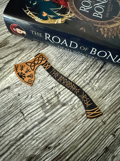 Decorative viking axe bookmark with runic designs on a book titled 'The Road of Bones' by Demi Winters.