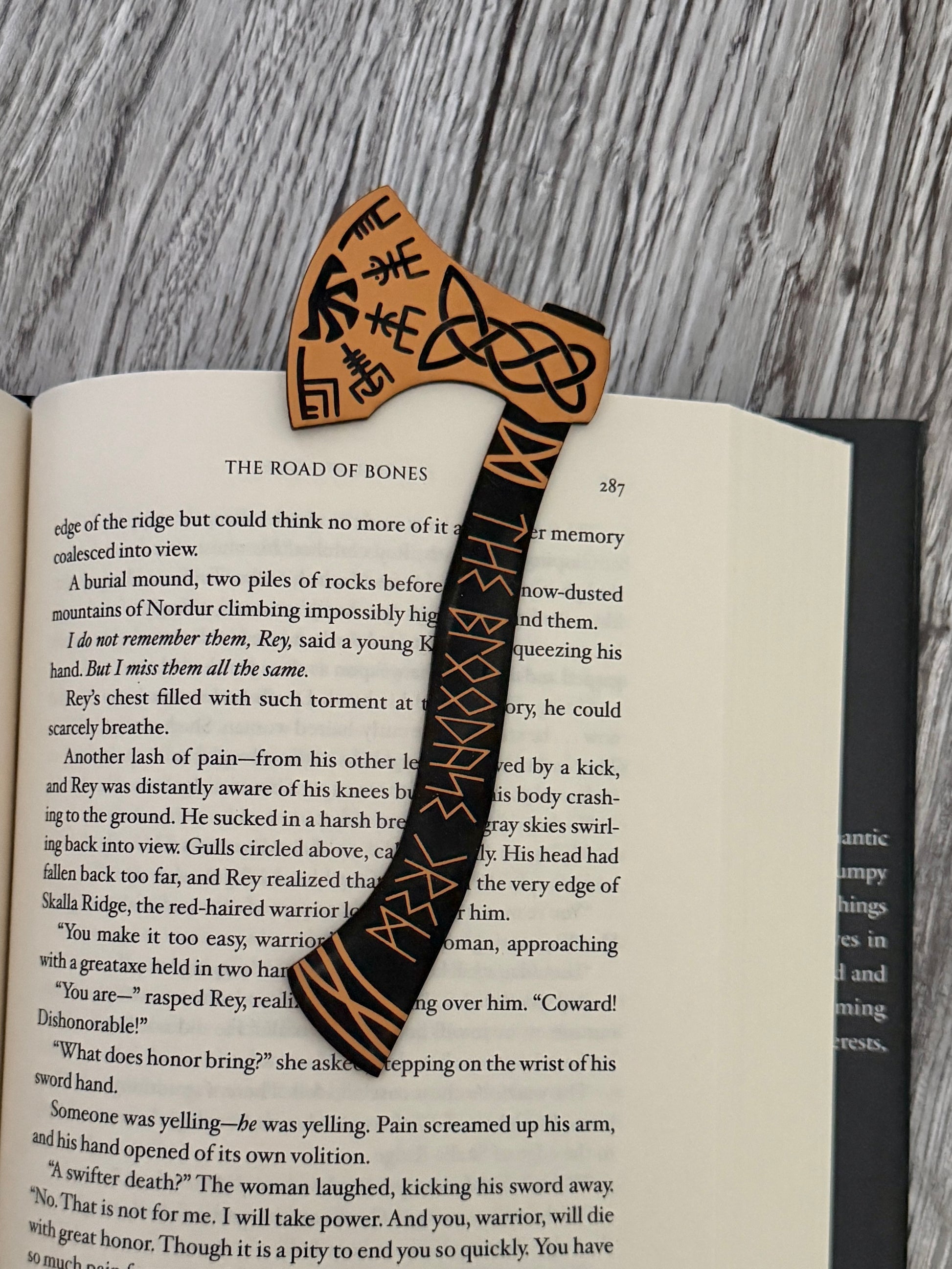 Acrylic engraved bookmark shaped like a viking axe with celtic knots and runes on an open book of The Road of Bones by Demi Winters.