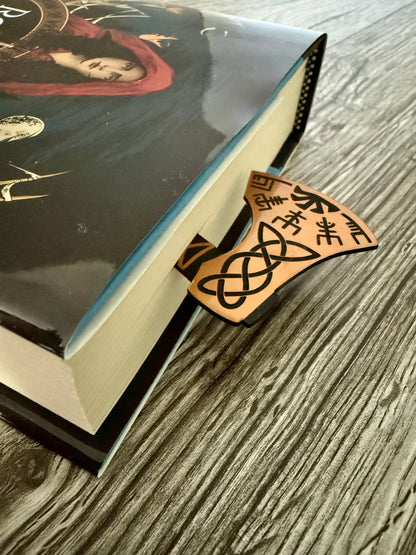 The Road of Bones book by Demi Winters with an axe bookmark sticking out the top on a wooden surface