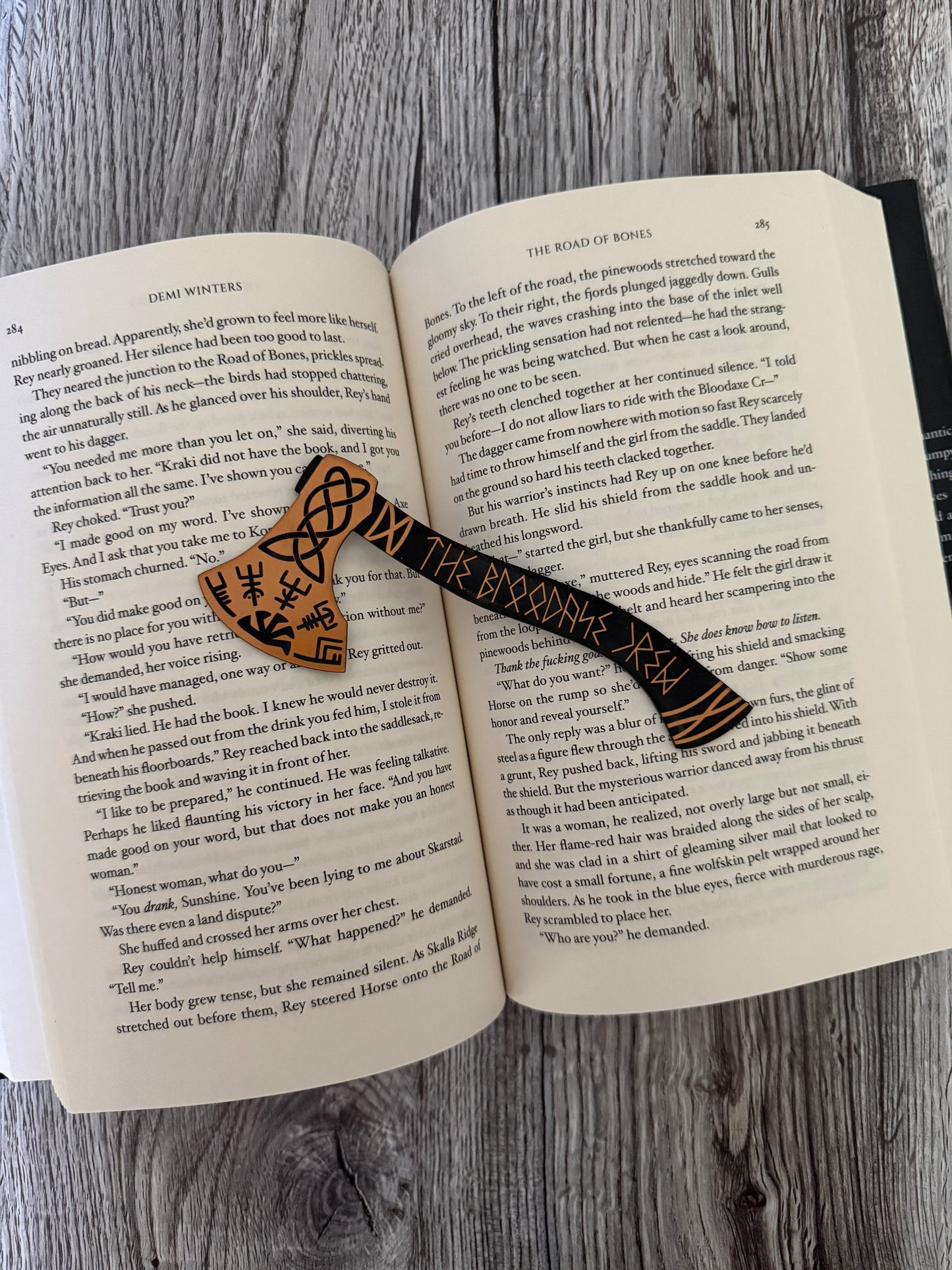 Open book 'The Road of Bones' by Demi Winters with a decorative axe bookmark on a wooden surface