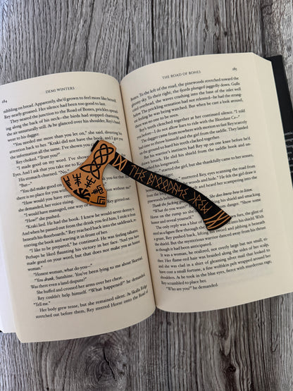 Open book 'The Road of Bones' by Demi Winters with a decorative axe bookmark on a wooden surface