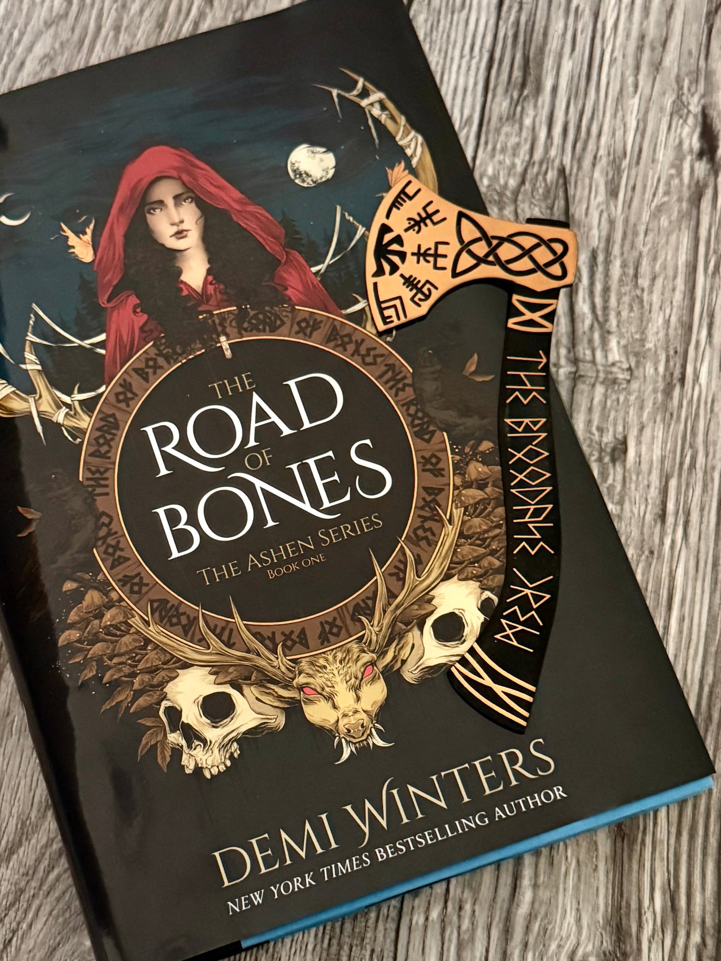 A Bloodaxe Crew axe-shaped bookmark sitting on top of 'The Road of Bones' book by Demi Winters on a wooden surface