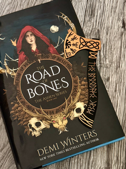 A Bloodaxe Crew axe-shaped bookmark sitting on top of 'The Road of Bones' book by Demi Winters on a wooden surface