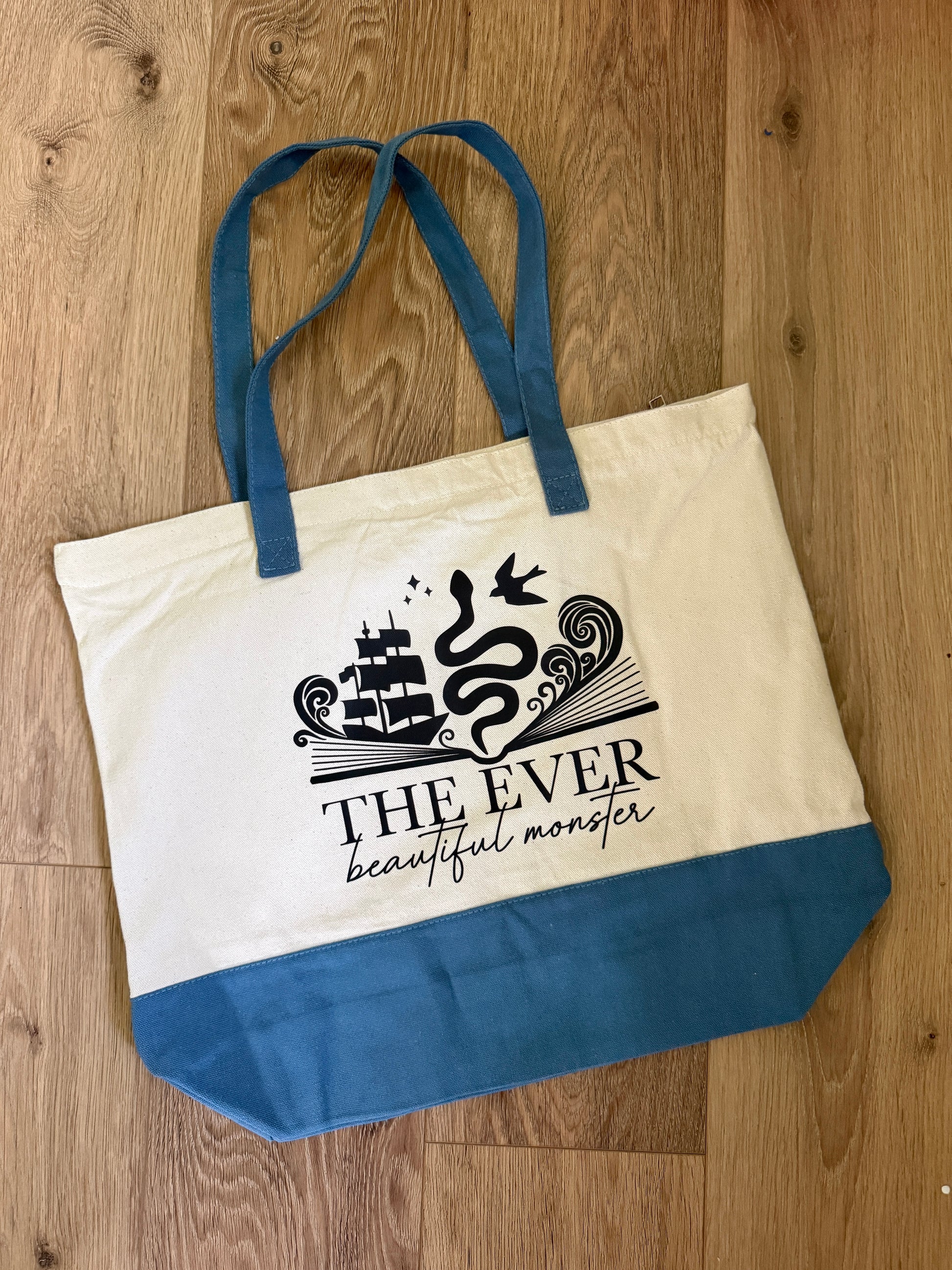 Tote bag with blue handles and bottom, featuring an officially licensed Ever King by LJ Andrews design with ship, serpent, songbird, waves and text 'The Ever: beautiful monster' on a wooden surface.