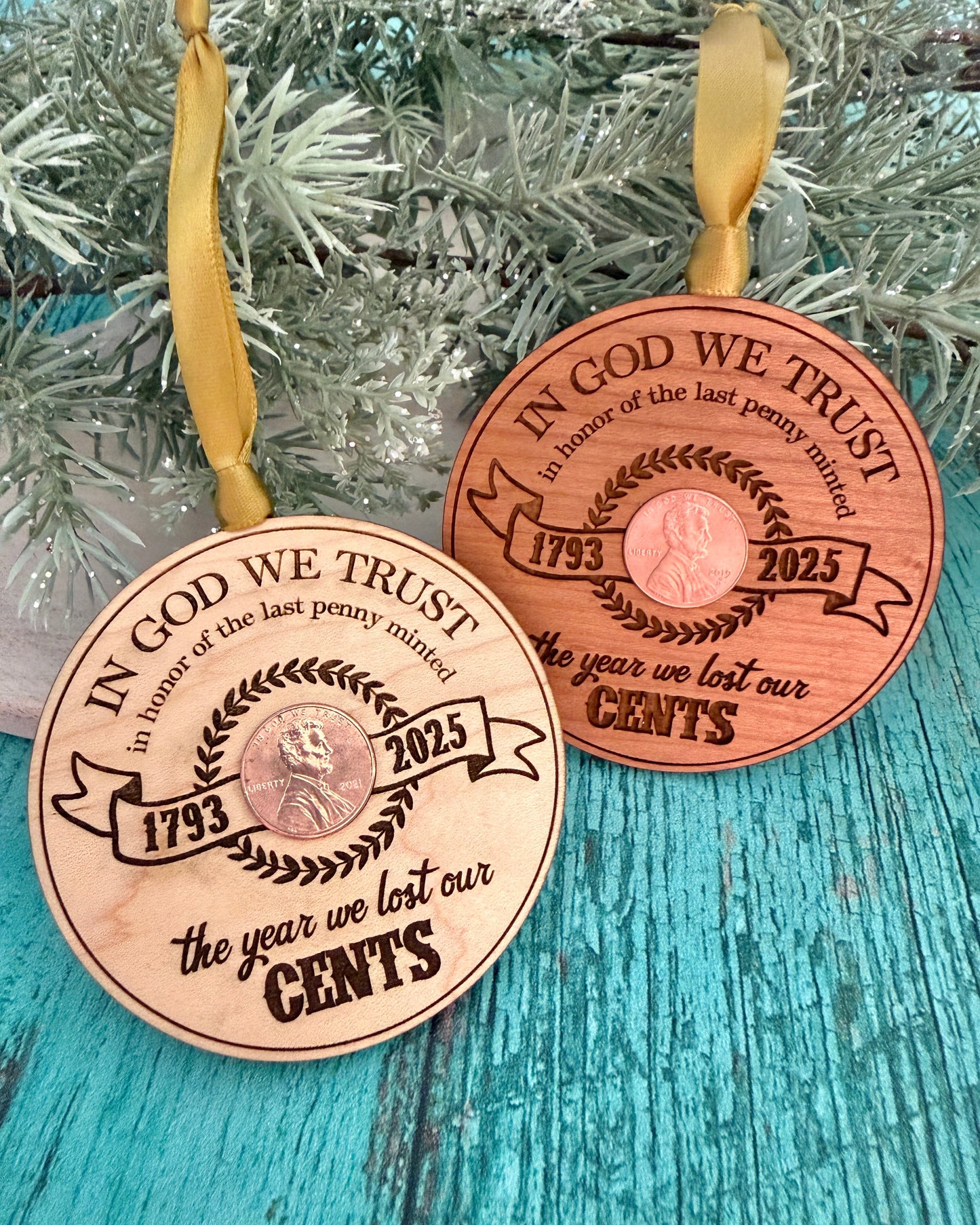 Two wooden ornaments embedded with pennies with engraved text "the year we lost our cents" on a teal surface with a Christmas tree background.