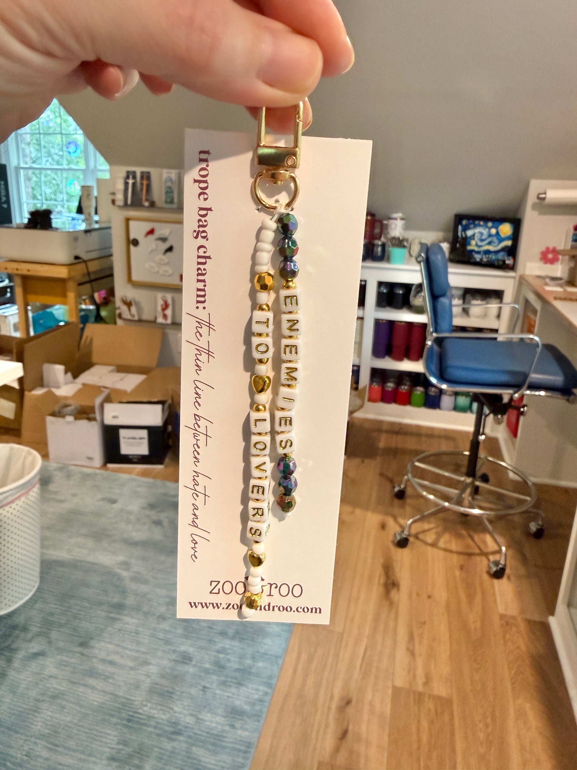 A hand holds a beaded keychain charm on a white zoo&roo display card in a home office or craft room. The keychain has gold-toned metal rings and beads, including white, iridescent black and gold beads, with the words 'ENEMIES TO LOVERS' spelled out in beads.