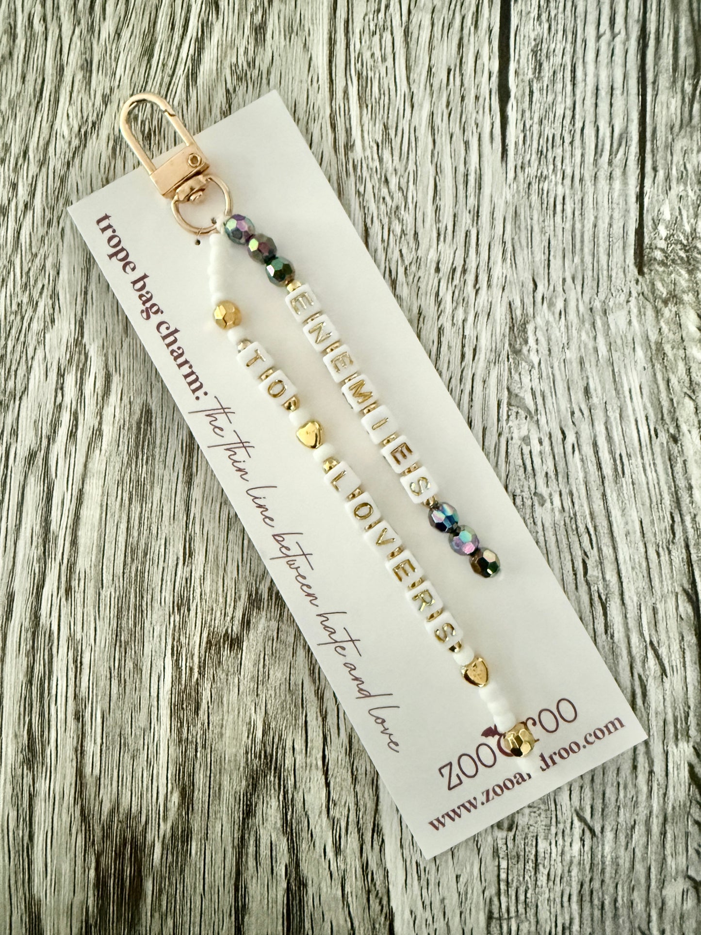 A beaded bag charm or keychain with gold-toned metal hardware and iridescent beads, that spell out 'ENEMIES TO LOVERS' attached to a white zoo&roo branded display card that reads 'trope bag charm: the thin line between hate and love,' and photographed on a weathered wood surface.