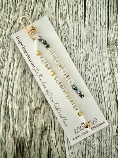 A beaded bag charm or keychain with gold-toned metal hardware and iridescent beads, that spell out 'ENEMIES TO LOVERS' attached to a white zoo&roo branded display card that reads 'trope bag charm: the thin line between hate and love,' and photographed on a weathered wood surface.