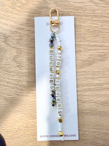A beaded keychain charm with gold-toned metal hardware, iridescent black, white and gold beads, with 'ENEMIES TO LOVERS' spelled out in white and gold letter beads, displayed on a white zoo&roo branded card over a light wood surface.
