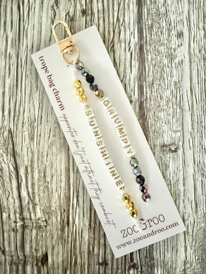 A gold-tone and multicolor beaded bag charm attached to a white zoo&roo branded display card, photographed on a rustic weathered wood surface. The card reads 'TROPE BAG CHARM' and the letter beads spell out 'GRUMPY' and 'SUNSHINE.'