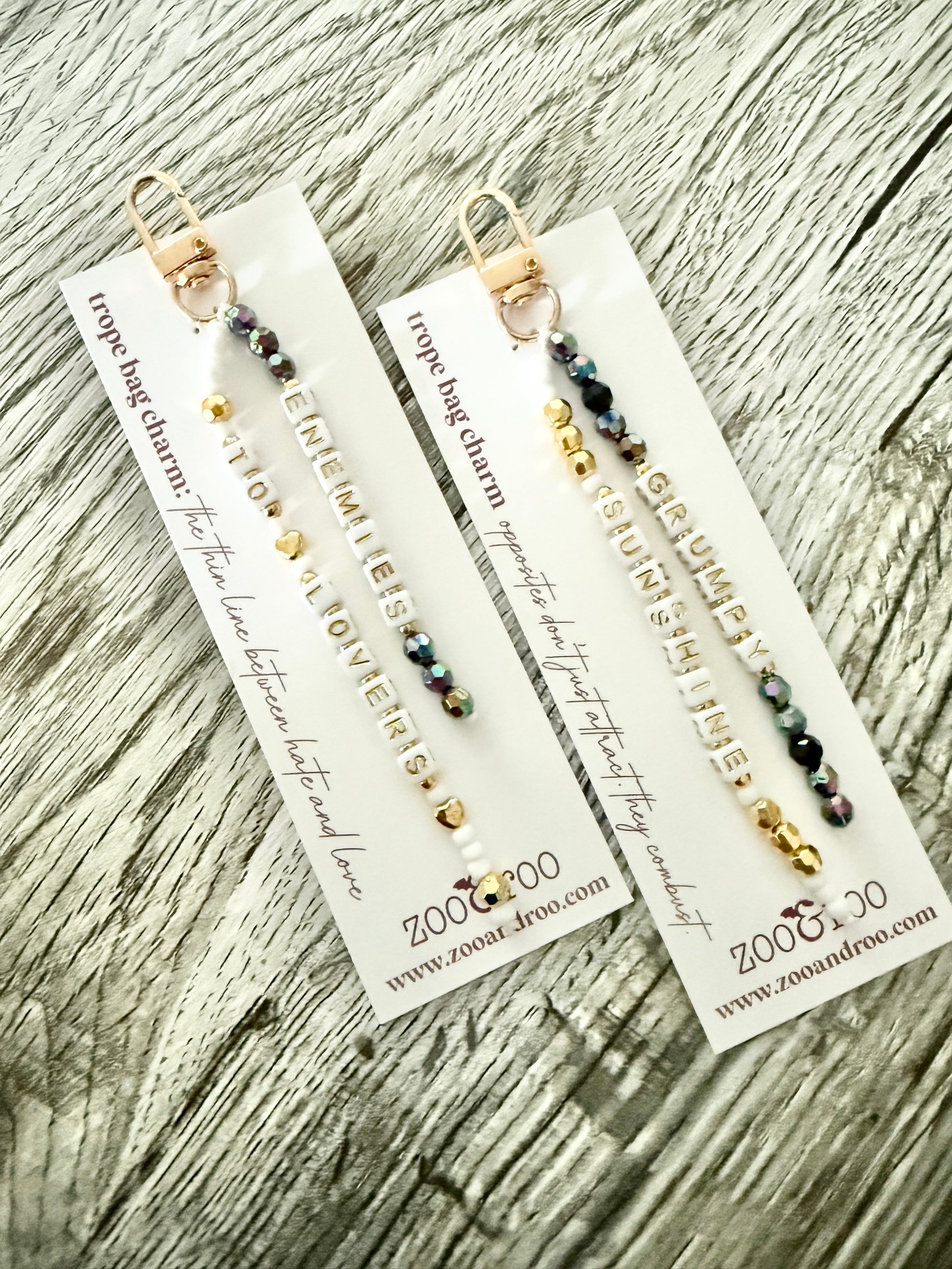 Two gold-toned bag charms with iridescent black, white and gold beads with letter beads that spell 'grumpy/sunshine' and 'enemies to lovers,' displayed on white branded zoo&roo cards over a weathered wood surface.