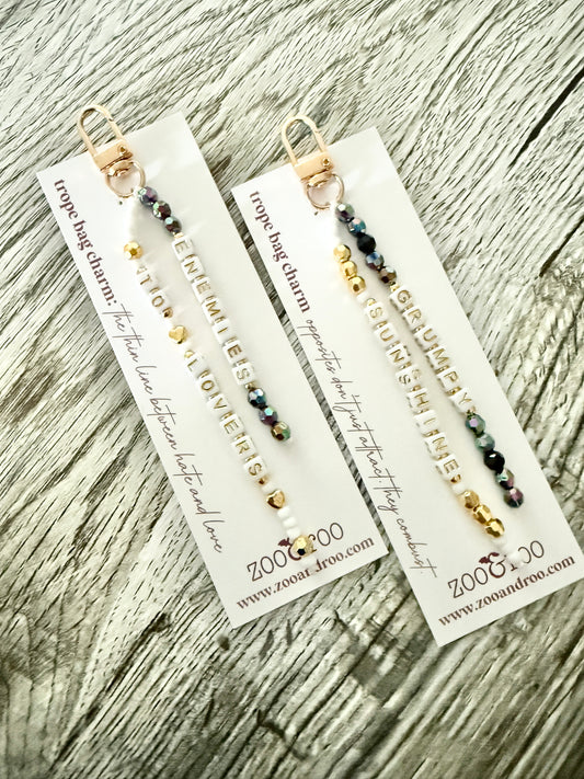 Two gold-toned bag charms with iridescent black, white and gold beads with letter beads that spell 'grumpy/sunshine' and 'enemies to lovers,' displayed on white branded zoo&roo cards over a weathered wood surface.