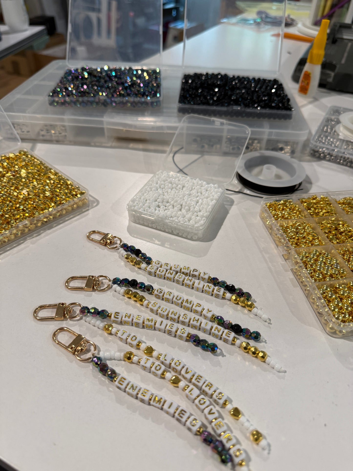 Several beaded keychains with gold-toned clasp closures and white letter beads spelling 'ENEMIES TO LOVERS' and 'GRUMPY SUNSHINE' are arranged on a white table, surrounded by trays of loose beads and spools of thread in a craft workspace.