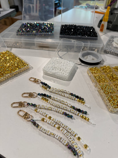 Several beaded keychains with gold-toned clasp closures and white letter beads spelling 'ENEMIES TO LOVERS' and 'GRUMPY SUNSHINE' are arranged on a white table, surrounded by trays of loose beads and spools of thread in a craft workspace.