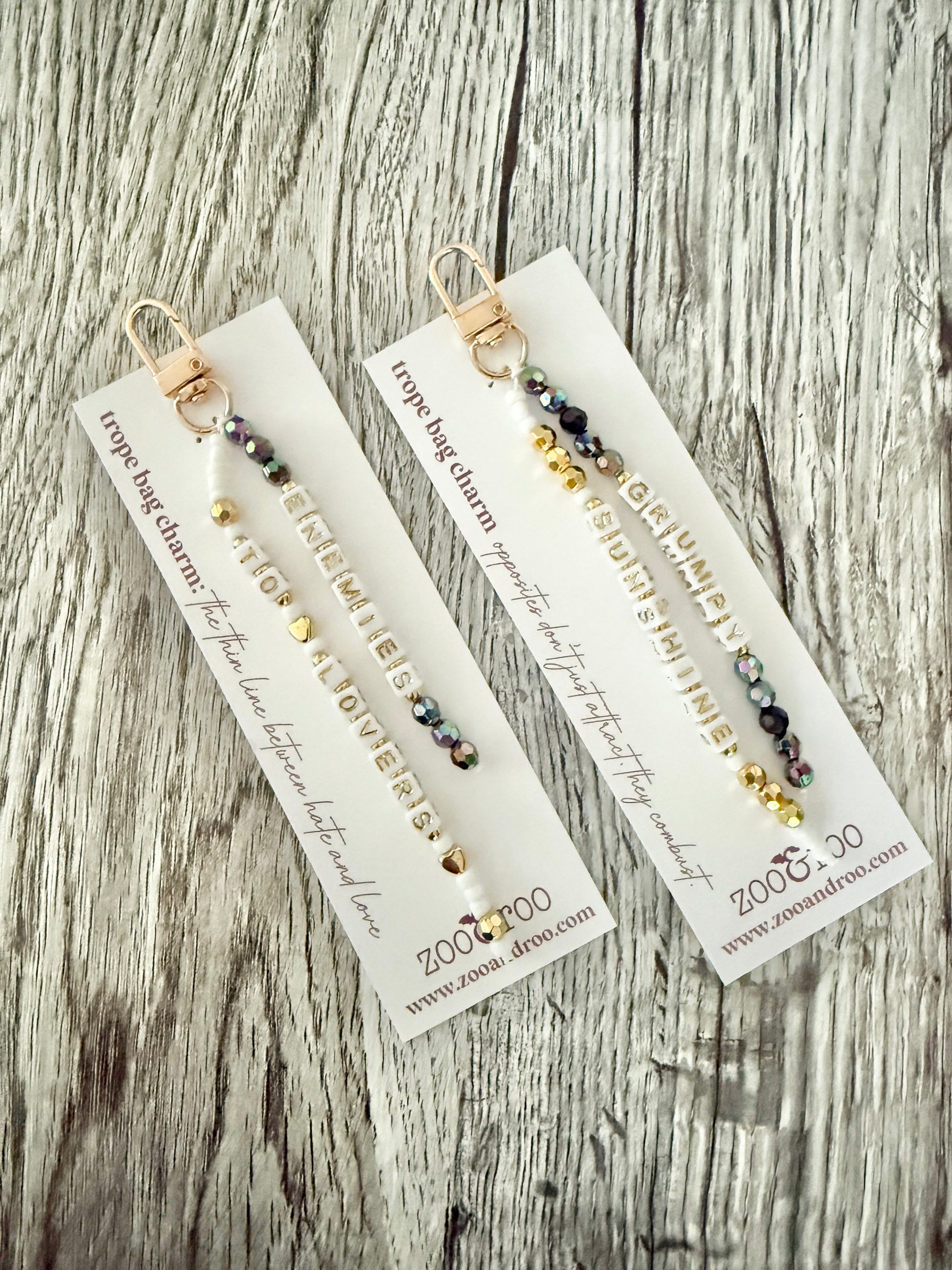 Two gold-toned bag charms with iridescent black, white and gold beads with letter beads that spell 'grumpy/sunshine' and 'enemies to lovers,' displayed on white branded zoo&roo cards over a weathered wood surface.